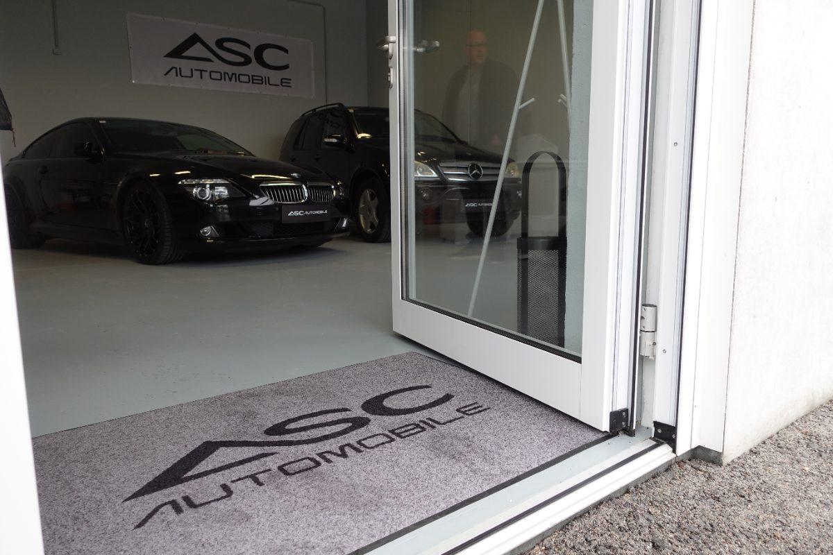Offene Glastür zum Showroom von ASC Automobile mit ASC-Fußmatte, dahinter zwei schwarze Fahrzeuge