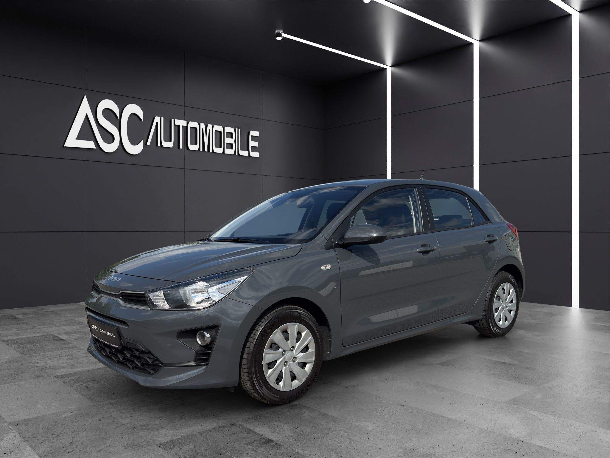 KIA Rio in Dunkelgrau, schräg von vorne links im Showroom, Fünftürer mit silbernen Radkappen